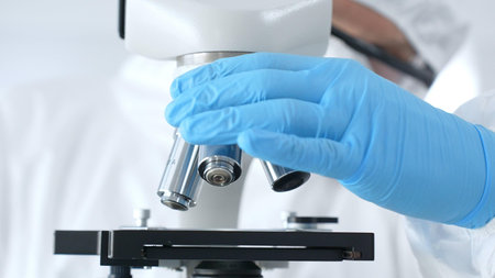 Close up of virologists hands in blue gloves adjusting microscope in laboratory, conducting...
