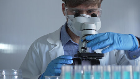 Scientist man, wearing a lab coat, mask, blue gloves and safety glasses, is using a binocular...