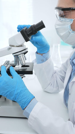 Female scientist analyzing sample through microscope, wearing protective lab gear during medical...