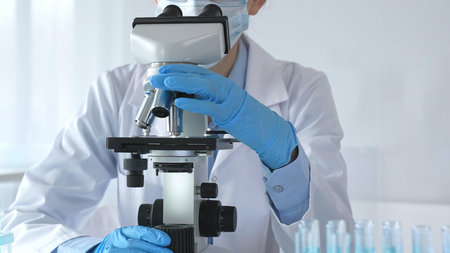 Female scientist wearing protective gear carefully adjusting microscope lens, analyzing medical...