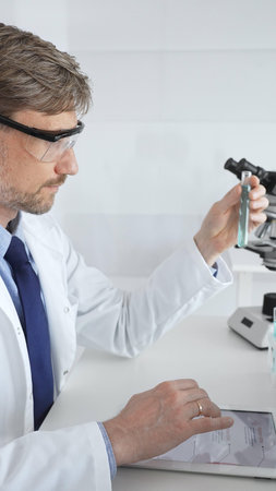 Male researcher in protective glasses holding test tube with blue liquid and working with tablet in...