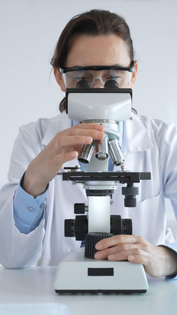 Female scientist wearing lab coat and protective glasses using microscope, analyzing samples in test...