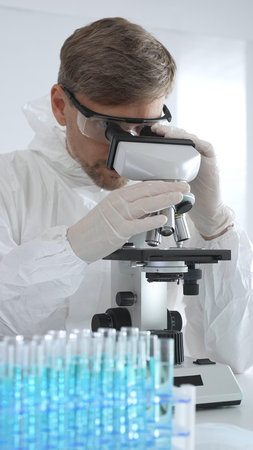 Male virologist in medical gown and gloves examines virus samples under microscope while conducting...