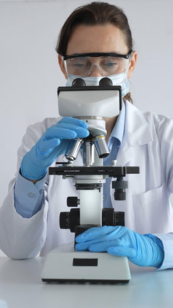 Female scientist examining microscope slide, wearing protective gear in laboratory, researching...