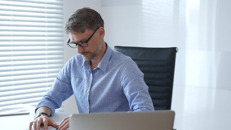 Businessman wearing blue sleek shirt and black glasses is concentrating while working at the desk in...