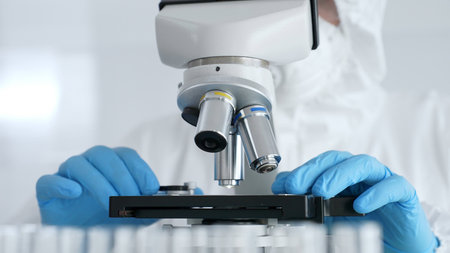 Scientific researcher or virologist wearing protective gear is tuning microscope lenses opposite the...