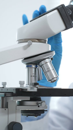 Close up of blue gloved scientists hand adjusting microscope in modern laboratory setting. Medicine,...