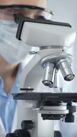 Scientist wearing lab coat and protective glasses using a modern microscope in a laboratory setting,...