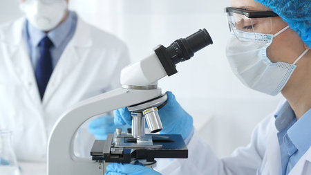 Close up of woman scientist wearing blue protective gear while examining specimen under microscope,...