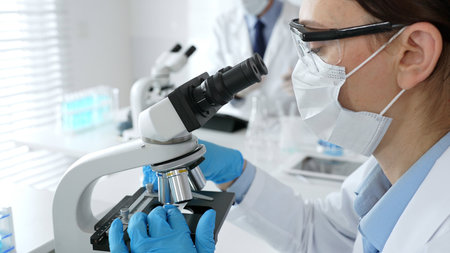 A female scientist in a blue mask and gloves is using a microscope and conducting scientific...