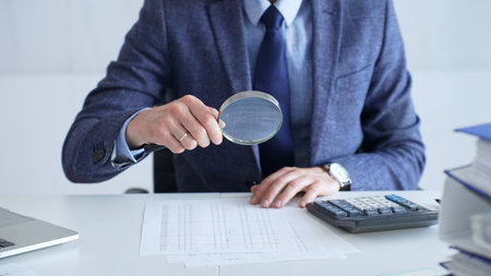Professional businessman examining financial documents, magnifying glass in hand, calculator...