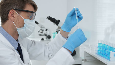 Masked male scientist in lab coat, protective glasses and blue gloves is adding reagent to test tube...