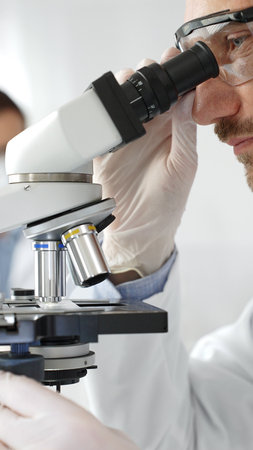 Close up of male researcher examining sample through microscope, wearing safety eyewear in sterile...