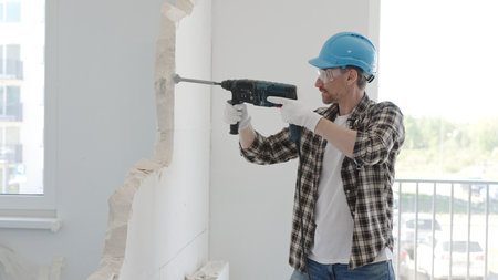 Man construction worker demolishing a wall with a hammer drill in protective gear, creating...