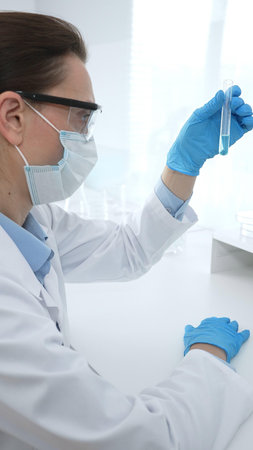 Female researcher examining clear liquid in test tube, sitting near microscopes and lab equipment...