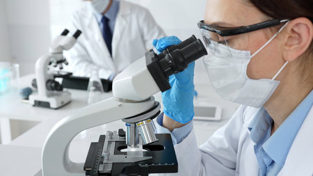 A female scientist in a blue mask and gloves is using a microscope and conducting scientific...