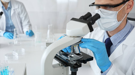 Masked scientist analyzing sample under microscope, colleague researching nearby in sterile medical...
