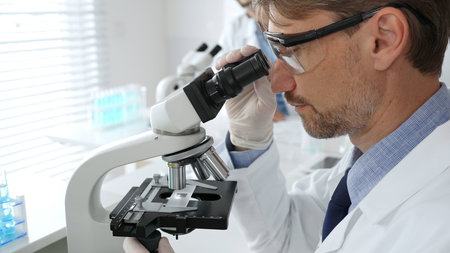 Medical scientist wearing gloves and protective glasses examining samples under a microscope in a...
