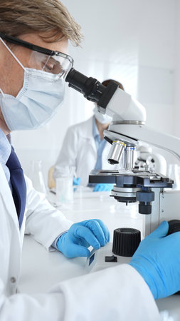 Masked male scientist analyzing sample under microscope, colleague researching nearby in sterile...