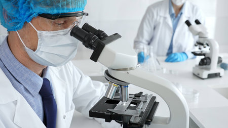 Senior male scientist wearing a protective mask, cap, and gloves, examining samples under a...