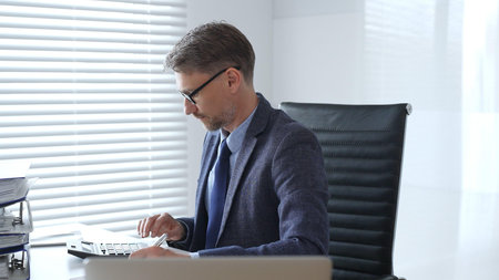 Mature entrepreneur in suit, tie and glasses is sitting at desk, writing notes and using calculator...