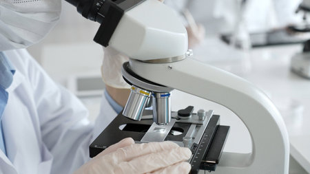 Gloved hands adjusting a microscope in a bright laboratory, emphasizing precision and focus in...