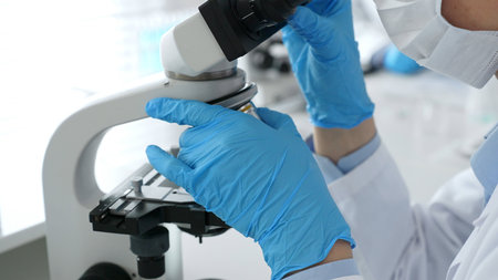 Scientific professional wearing blue protective gear examining microscope lens, performing detailed...