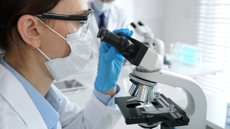 Female scientist wearing mask and gloves using microscope doing scientific research in modern...