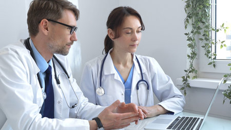 Two doctors are studying medical charts on a laptop, discussing patient data in a hospital or...