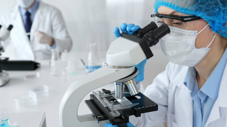 Female scientist wearing a mask and blue safety gear, using a microscope for research while a male...