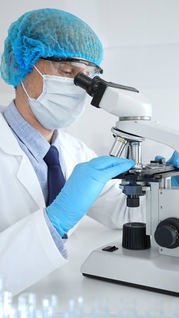 Senior male scientist wearing a protective mask, cap, and gloves, examining samples under a...