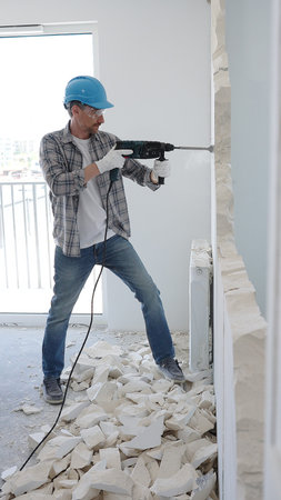 Male construction worker wearing grey checked shirt and blue hardhat is demolishing a wall with a...