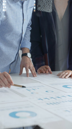 Business people are examining financial data, pointing at charts and graphs spread out on a table...