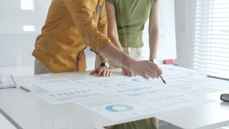 Two business professionals are examining financial reports, using a pen to point at specific data...