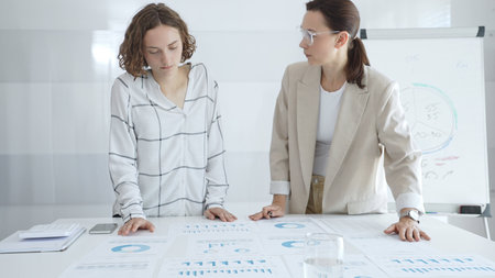 Professional businesswomen analyzing financial charts, collaborating in sleek corporate workspace,...