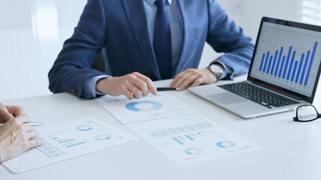 Business colleagues or partners are discussing financial data on a laptop and printed documents...