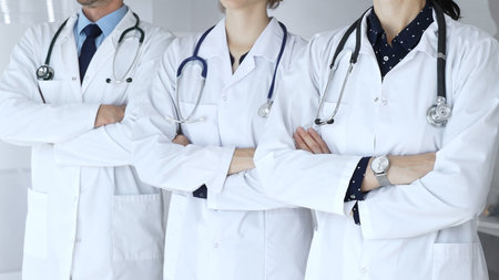 Doctors from a medical team are standing with arms crossed, wearing white coats and stethoscopes,...