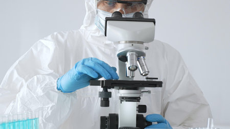 Male doctor in protective suit and gloves using microscope while analyzing samples while conducting research in sterile laboratory. Close-up of test tubes with blue liquidの写真素材