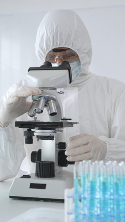 A medical virologist in protective gear examines viral samples under a microscope while conducting...