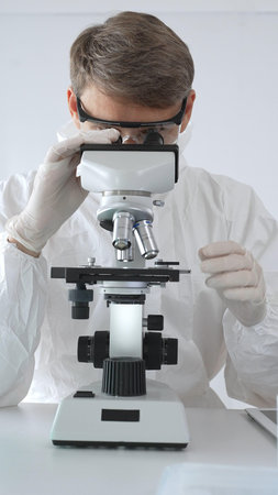 A male medical virologist in a medical gown and gloves examines viral samples under a microscope...