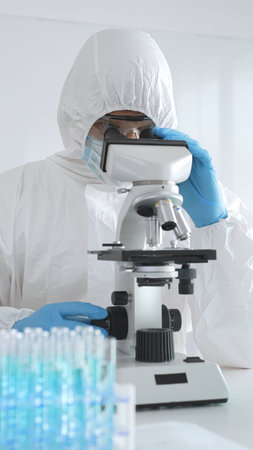 Medical scientist wearing protective suit using microscope while analyzing samples, conducting...