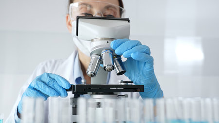 Woman science researcher wearing protective gear is checking microscope, analyzing blue test tubes...