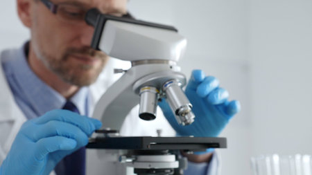 Close up of male scientist in white coat and blue gloves adjusting microscope, conducting...