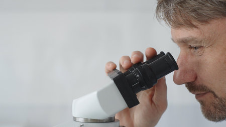 Close up of a man scientist carefully observing through a microscope in a brightly lit laboratory,...