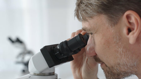 Close up of a man scientist carefully observing through a microscope in a brightly lit laboratory, conducting research for developing new medicines and treatments. Medicine and science conceptの写真素材