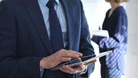 Businessman in a blue suit using a tablet during a corporate meeting, interacting with the touchscreen as blurred colleagues collaborate in the background, in office. Business people conceptの写真素材