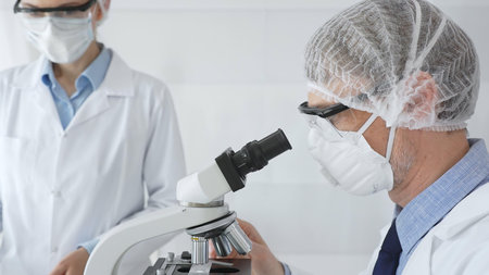 Senior male scientist wearing protective mask, glasses and hairnet using microscope with young female colleague assisting, holding tablet and taking notes in laboratory. Medicine and science conceptの写真素材