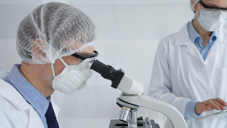 Senior male scientist wearing protective mask, glasses and hairnet using microscope with young...