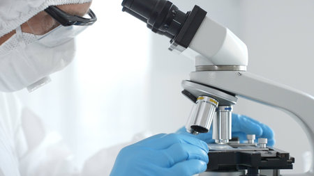 Medical virologist in a hazmat suit, mask, and blue gloves, diligently working at a microscope in a...
