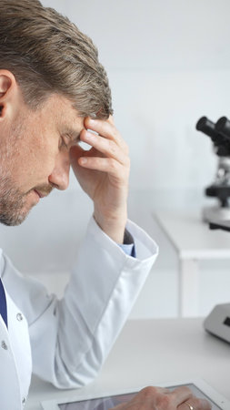 Confident male mature scientist is thinking and analyzing data on a tablet in his laboratory, with a microscope in the background, suggesting advanced scientific research. Medicine and science conceptの写真素材